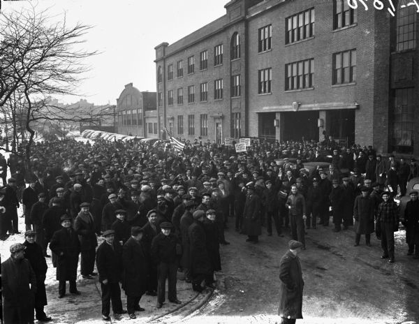 Elevated view of striking International Harvester factory workers holding signs and an American flag as they stand near the company's Tractor Works.