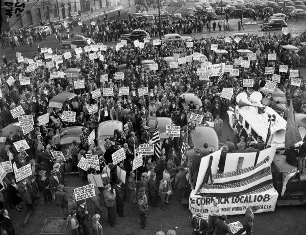 Elevated view of members of the Farm Equipment Workers Union meeting in a parking lot of the McCormick Works factory located at the corner of Oakley and Blue Island Streets. Individuals hold picketing signs, and a sign attached to a truck bed in the foreground reads, "McCormick Local 108."