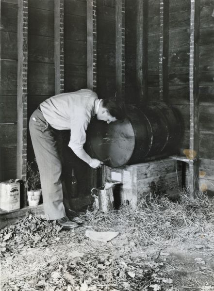 A man is filling a metal pitcher with oil from a barrel stored in a garage with litter on the ground, illustrating a farm hazard.