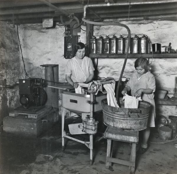 A woman and a girl are using an electric washing machine to clean clothing or bedsheets in a basement. The machine is powered by a DELCO-Light Plant system, with the generator in the corner of the room ,and glass batteries lined up on wall-mounted wooden shelving.
