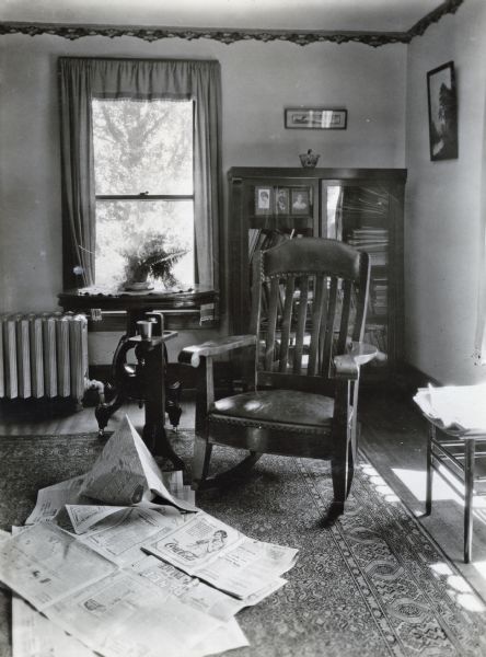 Pages from a newspaper are scattered on a rug at the base of a rocking chair in a farmhouse living room. A radiator and bookcase are behind the chair.