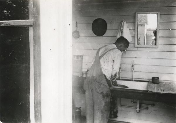 A man wearing overalls washing his hands in a sink in a farmhouse washroom. There is a reflection of a man in the mirror over the sink, who appears to be using a camera to take the photograph.