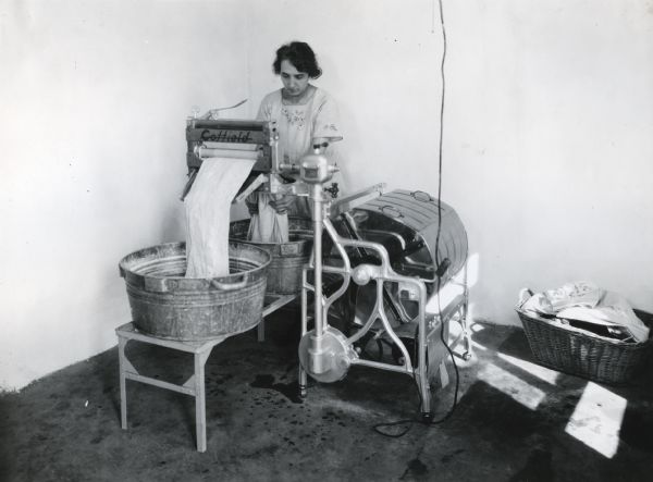 Mrs. Waggoner using a Coffield electric washing machine and wringer to launder clothing on International Harvester's Hinsdale experimental farm (Harvester Farm). A laundry basket is sitting on the ground beside her.