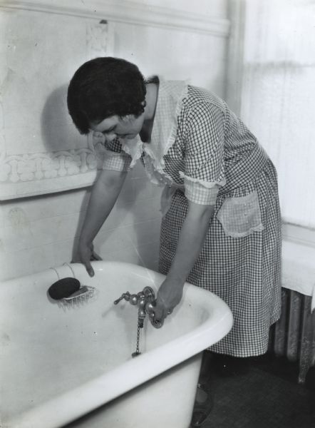 A woman wearing a checkered dress is turning the faucet on a bathtub at International Harvester's Hinsdale experimental farm (Harvester Farm).