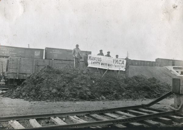 Men hold a sign that says "Car No. 1 Manure Secured by Y.M.C.A. for Eighth Ward War Gardens" while standing on top of a manure pile in a railroad yard. 
