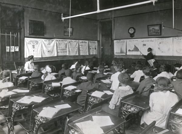 A group of students and a teacher are sitting at desks in a classroom while listening to a lesson on the dangers of house flies. Agricultural lecture charts about flies are hanging on the chalkboards on the walls of the room.