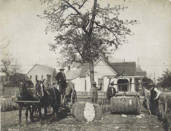 Men in a residential backyard using a hanging scale to weigh bales of cotton loaded on a wagon pulled by a team of work horses.