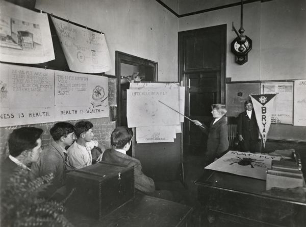A student is using a pointer to direct the attention of seated students toward a poster labeled "Life Cycle of a Fly" during a lesson on the dangers of house flies. Additional posters on the subject of flies and health are hanging around the room. A boy holding a banner labeled "Bryant" is standing in the background.