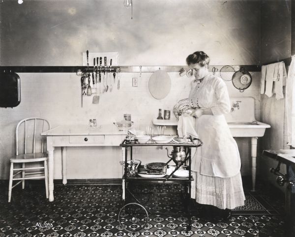 A woman wearing an apron is standing near a wheeled butler's tray while drying dishes in a kitchen. A sink, table, chair and utensils are behind her.