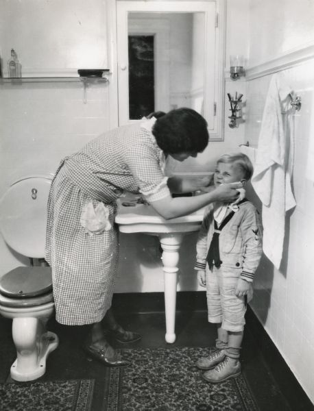A woman is washing a boy's face with a washcloth in a bathroom on International Harvester's Hinsdale experimental farm. The boy is wearing a sailor suit with what appears to be a United States Naval insignia on the sleeve.