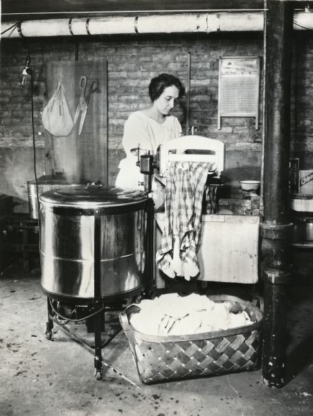 Mrs. J.E. Waggoner passing plaid fabric through a wringer after using an electric washing machine. A washboard is hanging on the wall in the background, and a laundry basket is on the floor.