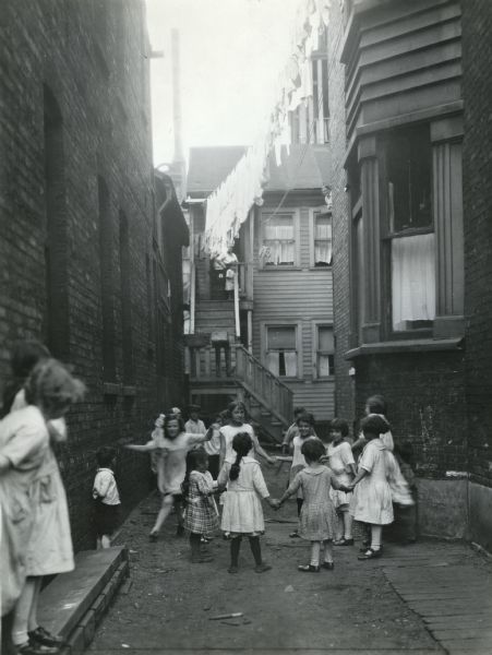 Children joining hands while playing in an alleyway between brick buildings in an area of tenement housing. A line of laundry suspended over the alley is hanging above them.