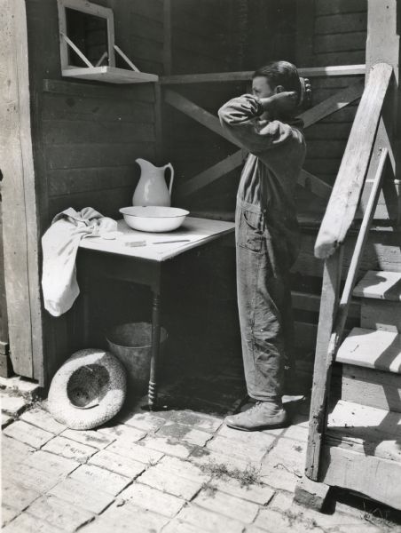 A boy using a pitcher and bowl set on a table outdoors to wash up after doing farm chores.
