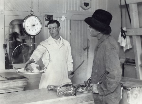 A butcher holds a knife as he weighs a piece of meat on a hanging scale. A male customer wearing a one-piece work suit and hat waits on the opposite side of the counter.