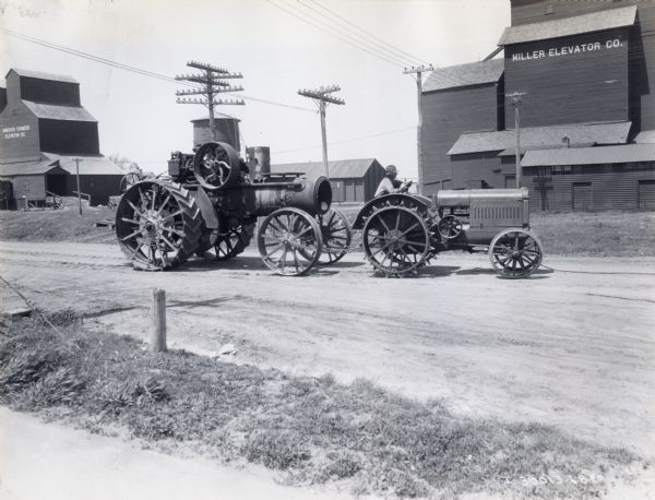A steam tractor is hauled along a dirt road by a McCormick-Deering 10-20(?) tractor. A building with text reading "Miller Elevator Co." stands in the background.