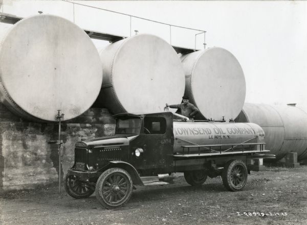 A man, standing in front of three large storage tanks, is filling the tank on the back of an International Model 63 truck owned by the Townsend Oil Company.