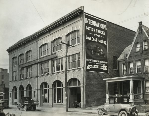 Exterior view of the International Harvester Company Parkersburg branch building. Automobiles and trucks are parked along the street running in front of the building, and a residential building stands beside it.