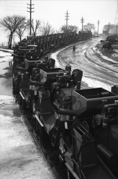 Elevated view of a line of combines loaded on open railroad cars on tracks on one side of a wet and snowy dirt road at International Harvester's East Moline Works (factory).