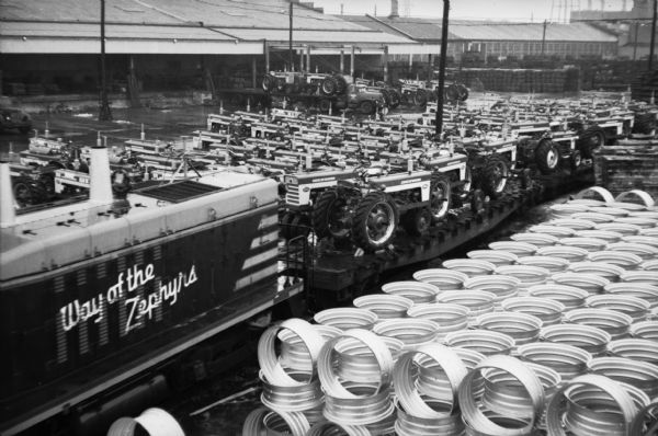 Elevated view of Farmall tractors are loaded onto railroad cars as they prepare to leave International Harvester's Farmall Works factory. On the locomotive is painted a sign that reads: "Way of the Zephyrs". In the foreground are stacks of what appears to be tire rims.