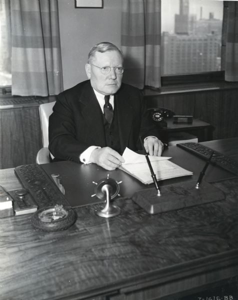 Portrait of C.R. Morrison, a Vice President of International Harvester Company, sitting at his desk. Windows behind the desk show a skyline.