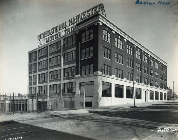 Exterior view of an International Harvester Company branch building. Large show windows line the first floor along the sidewalk. A large electric sign on the roof reads, "International Harvester Motor Trucks" and street railroad tracks run down the street. Residential houses are in the far background.