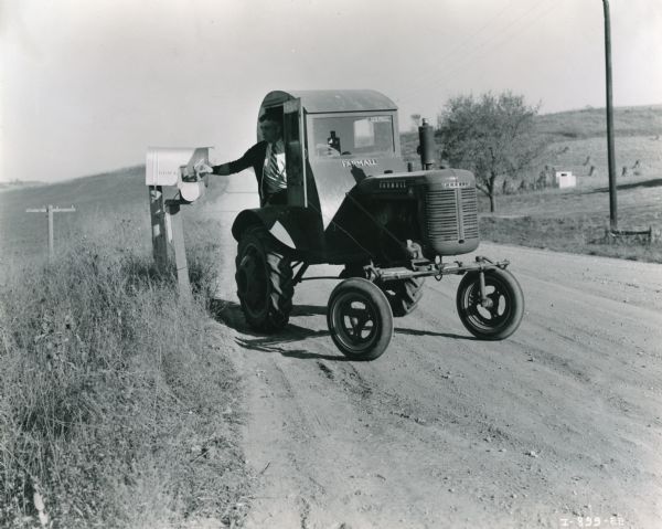 Francis Shirbroun delivers mail to a mailbox in a Farmall A tractor that has been fitted with a homemade cab for delivering mail. The cab has a "FARMALL" decal as well as a "U.S. MAIL" decal. The original caption reads: "Francis Shirbroun, Coon Rapids, Iowa delivers mail over a rural route of 51 miles with 195 boxes. When the roads are bad Farmall A covers it in 5 1/2 to 6 hours on not over 6 gallons of fuel."
