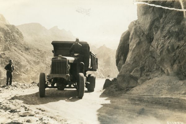 A man appears to be backing up an International truck through a dirt road pass in a mountainous area during the construction of Hoover Dam. He is standing in the driver's seat holding onto the steering wheel and facing towards the back of the dump truck. Another man stands to the left at the edge of the road.