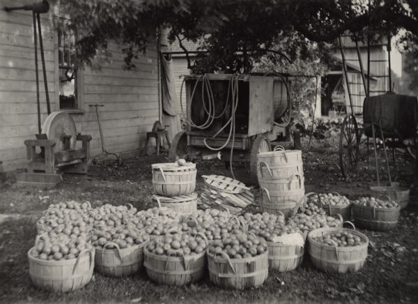 Apples in baskets in front of a "farm orcharding sprayer" at R.R. Robertson's. Farm buildings are in the background. On the left is a grinding wheel powered by a belt attached to an axle protruding from the side of a building.