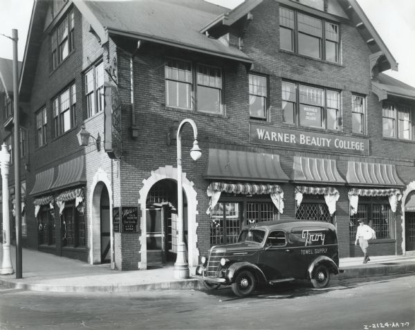 View across street of an International D-2 panel truck owned by the Troy Towel Supply Company parked outside Warner Beauty College. A man is walking on the sidewalk with a bundle towards the truck.