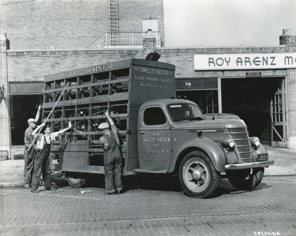 Three men load a piece of sheet glass onto the back of an International D-35 truck owned by the Dwelle — Kaiser Company. Text on the truck reads: "The Dwelle — Kaiser Co. Glass — Mirrors — Paints. Buffalo, N.Y." The service entrance to a building labeled "Roy Arenz" is in the background.