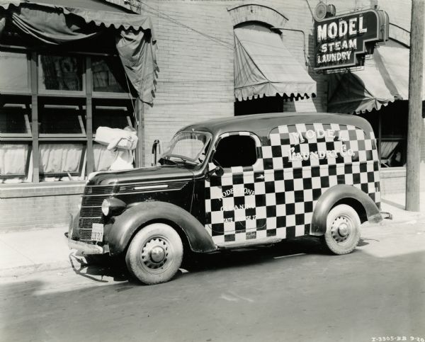 An International Model D-2 truck owned by Model Laundry Company parked outside the business' storefront. The truck is painted in a checkerboard pattern and the text on its side reads: "Model - Tone Cleaning. State License 27. Model Laundry Co."