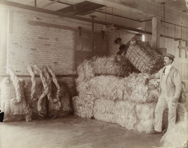 A man is standing in a warehouse beside bales of manila fiber used to make twine at the McCormick Twine Mill.