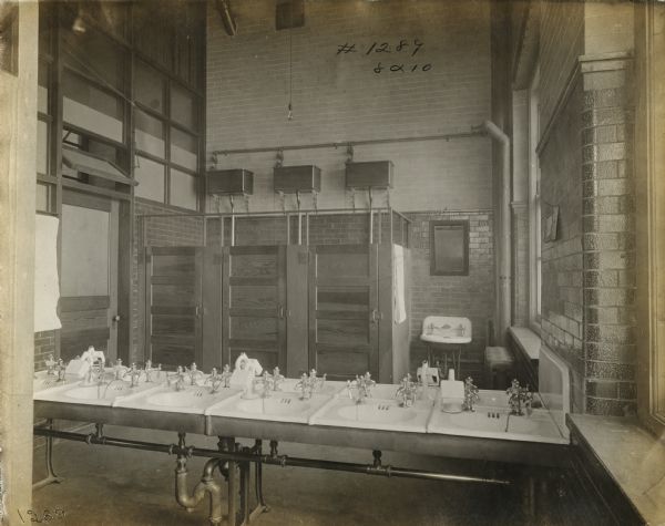 View of a bathroom at the McCormick factory. A row of sinks are standing in the foreground, and several toilet stalls with overhead tanks are along the back wall. There is a window on the right, and a door is on the left.