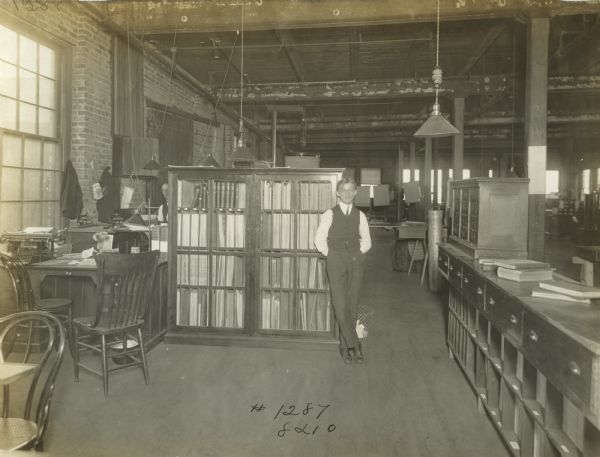 A man stands beside a bookcase in a factory office, possibly in the bookkeeping or accounting department, at the McCormick Works. Several desks and cabinets are arranged throughout the room. Another man is seated at left just behind the bookcase.