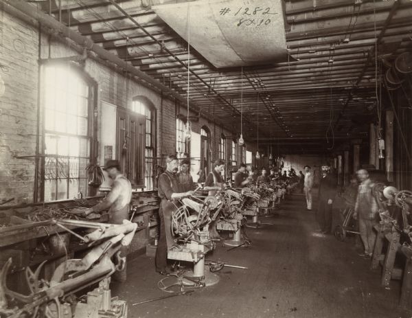 Factory workers using hammers and other tools to construct binder attachments(?) while standing at workstations inside a room at the McCormick Works. Piles of metal parts are arranged in boxes along the wall beneath windows.