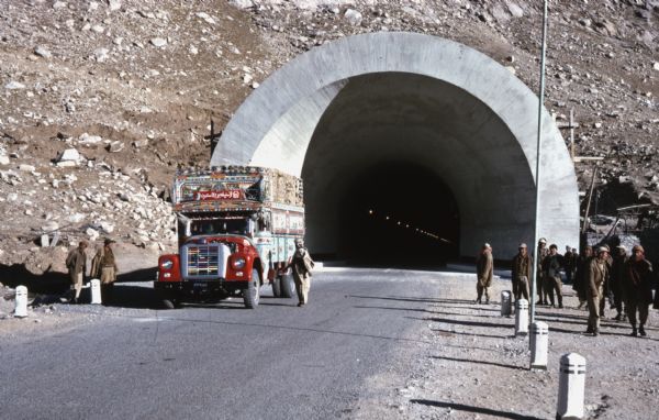 A colorfully painted and decorated International truck on a road in front of the arched entrance to a mountain tunnel in Afghanistan. Men stand near the truck on the left, and another group of men are on the right side of the road. The truck may be serving as a bus.