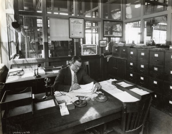 Factory Manager at Fort Wayne Works A man, likely a manager at International Harvester's Ft. Wayne Works, sits at a desk working on papers. There are phones, filing cabinets and other office equipment. A framed poster with the text: "Quality More Sales Steady Employment" is hanging on the wall in the background, along with a advertisement featuring an illustration of a zeppelin.