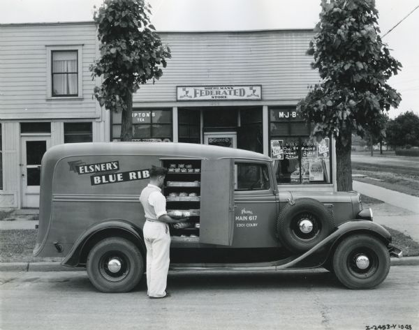 A man unloads loaves of bread from the side of an International C-1 truck owned by Elsner's Blue Ribbon Bakery. The bread is being delivered to Moehlman's Federated Store, which is behind the truck.