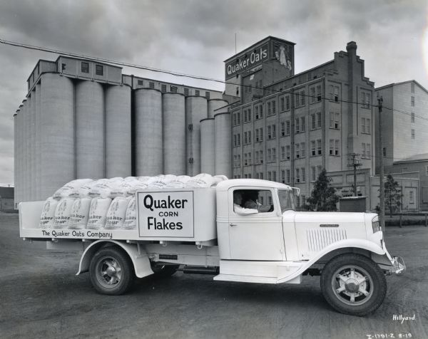 An International C-35 truck owned by the Quaker Oats Company parked in front of the Quaker Oats factory in Saskatoon, Saskatchewan, Canada. Sacks of flour are stacked in the open back of the truck, and two men sit inside the cab. Large storage tanks are next to the factory building in the background.
