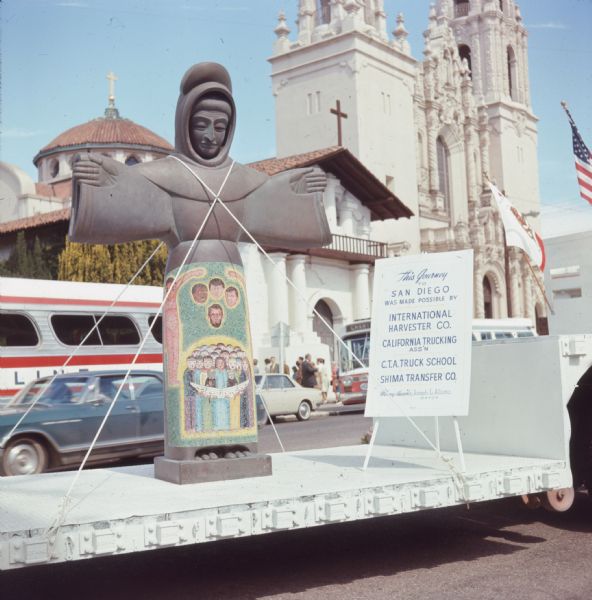 Color photo of the metal statue "St. Francis of the Guns" on a truck trailer parked on the street. The statue represents St. Francis of Assisi, was created by San Francisco sculptor Beniamino Bufano, and was cast from the melted down barrels of 1,900 guns. The statue has a mosaic of assassination victims President John F. Kennedy, Robbert Kennedy, Martin Luther King, and Abraham Lincoln, with a group of people singing below them on it's lower half. In the background are vehicles, a small group of people, and a California mission church. A placard on the trailer reads: "This journey to San Diego was made possible by International Harvester Co., California Trucking Ass'n, C.T.A. Truck School, Shima Transfer Co., many thanks Joseph L. Alioto, Mayor."
