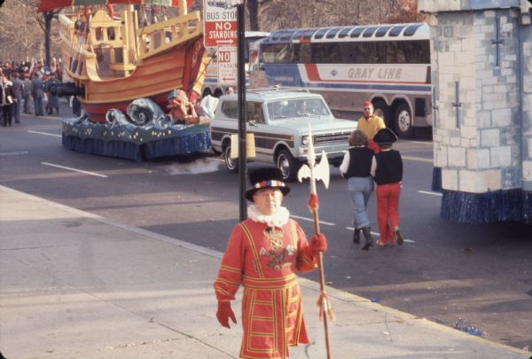 Thanksgiving Day Parade, either Macy's Parade in Manhattan, New York, or Gimbels' Parade in Philadelphia, Pennsylvania. A man in the foreground walking along the sidewalk is in a red Beefeater costume with yellow and black pinstriping carries a polearm. He has a coat of arms consisting of a crown above three flowers on his chest, red gloves, and a black hat with a band made of red, white, and blue squares. Behind him is a "Bus Stop, No Standing" and "No Parking" sign. A pirate ship float towed by a blue and brown International Harvester Scout 4 x 4 is coming along the street, and a bus with the words "Gray Line" on the side is in the opposite lane. Spectators can be seen in the background.