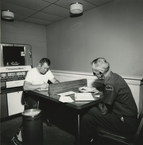 Two truck drivers filling out paperwork at a table in an American Oil Company truck stop. Behind them is a vending machine for cigarettes.