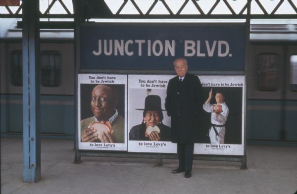 An older man is standing in front of three subway advertisements for Levy and Son's Bakery rye bread of New York City. The man is company president Samuel Rubin, who is wearing a knee-length coat over a suit. A sign behind him reads: "JUNCTION BLVD." and a subway train is in the background. The three advertisements all read: "You don't have to be Jewish to love Levy's real Jewish Rye." The photo caption reads: "Samuel Rubin, Levy's President, with three acclaimed subway ads. They depict entertainer Godfrey Cambridge, an American Indian, and a karate master."