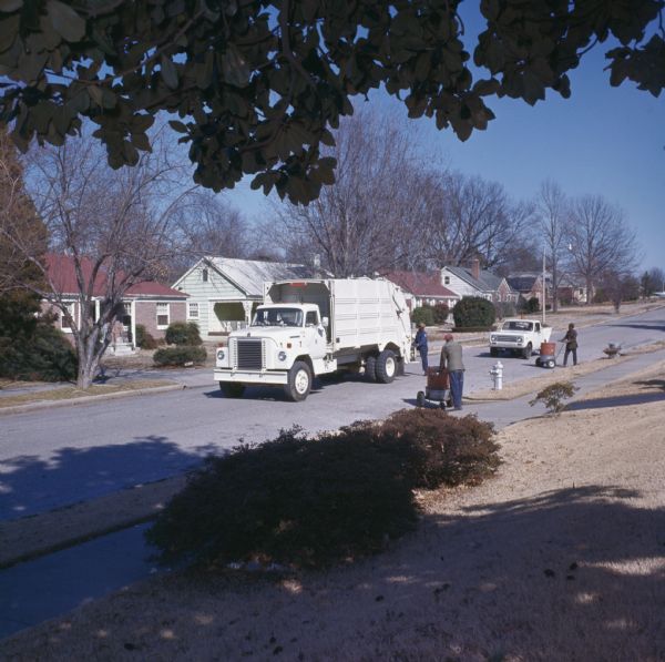 View from lawn in a residential neighborhood of three men pushing trash cans on hand carts towards a garbage truck. Behind the garbage truck is an International Harvester Scout with a dump body. The garbage truck is either an International Harvester model 1910A or 2010A.