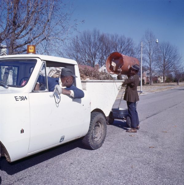 An African-American man is dumping a barrel of trash into an International Harvester Scout with a dump body driven by another African-American man. They are on a street in a residential neighborhood, and trees and houses can be seen in the background.