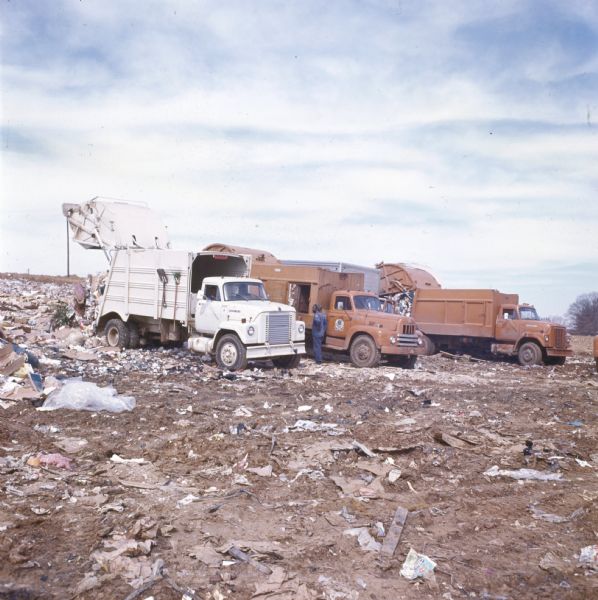 Three garbage trucks are pictured in a landfill dumping trash. An African-American man is standing by one of the trucks. The trucks are likely International Harvester model 1910A or 2010As. Various garbage and mud can be seen in the picture.