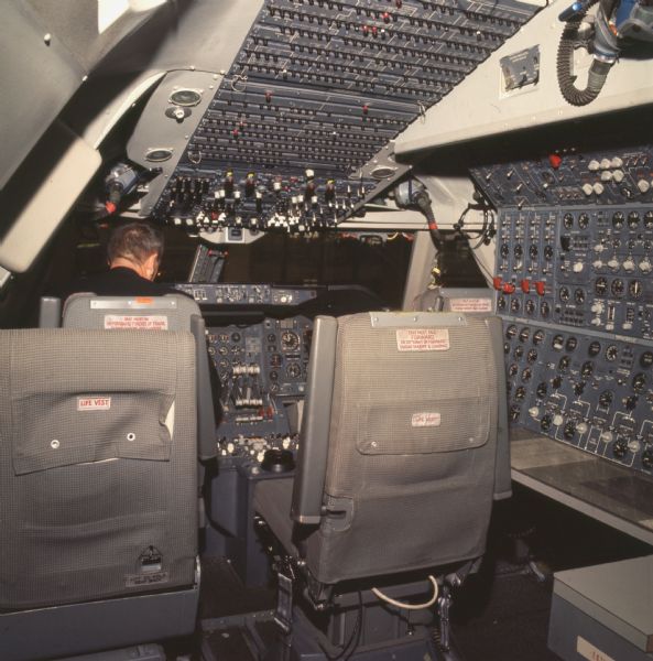 Rear view of man sitting in a Pan American World Airways Boeing 747 airplane cockpit. There are three seats and numerous switches, dials, knobs and indicators.