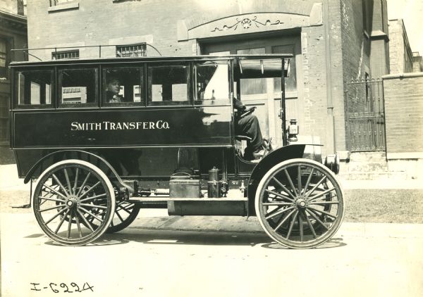 Side view of right side of an International bus used by the Smith Transfer Co. The front of the bus is open and a person can be partially seen sitting in the driver's seat. There is a luggage rack on the roof. A male passenger sits on the bus looking out the window. Behind the bus is a large brick building.