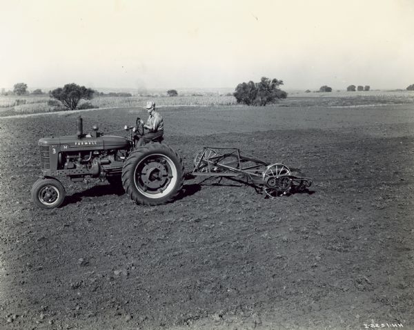 Farmall M Pulling 12-Foot No. 4 Rod Weeder Slightly elevated left side view of man driving a Farmall M tractor pulling 12-foot No. 4 rod weeder with regular rigid tractor hitch and screw-type regulator in a field.