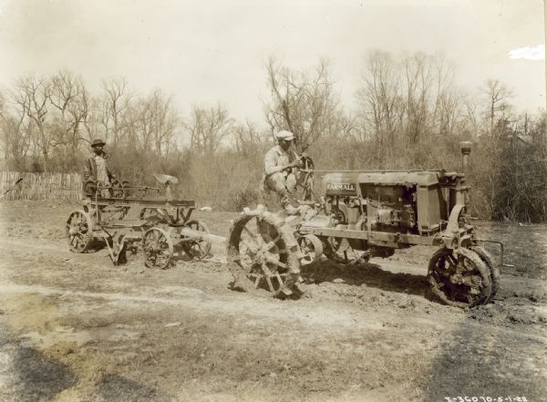 Right side view of two men in a wet, muddy field. One of the men is driving a Farmall tractor which is pulling another man who is standing on the back of a Russell "Junior." Oscar E. Wood.